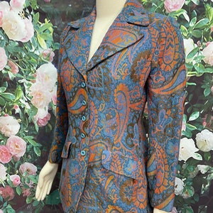 May include: A blue, orange and brown paisley patterned skirt suit. The jacket has a notched collar and a button closure. The skirt is knee-length and has a straight silhouette.