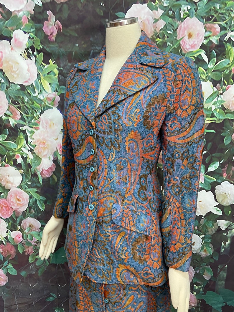May include: A blue, orange and brown paisley patterned skirt suit. The jacket has a notched collar and a button closure. The skirt is knee-length and has a straight silhouette.