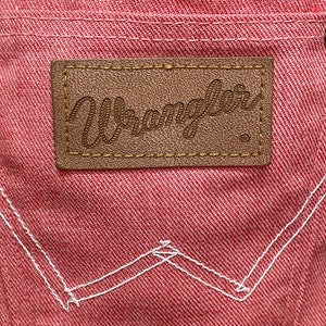 May include: A close-up of a pink denim fabric with a brown leather patch that says "Wrangler" in a cursive font. The patch has a white stitching around the edges. The denim has white stitching in a "W" shape.