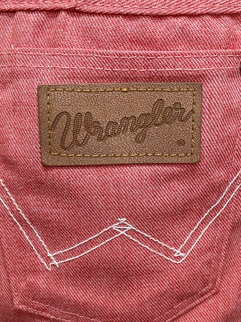 May include: A close-up of a pink denim fabric with a brown leather patch that says "Wrangler" in a cursive font. The patch has a white stitching around the edges. The denim has white stitching in a "W" shape.