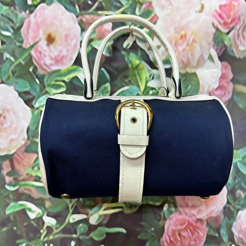 May include: A navy blue and white handbag with a white leather strap and gold buckle. The bag has a cylindrical shape and a white trim around the top and bottom.