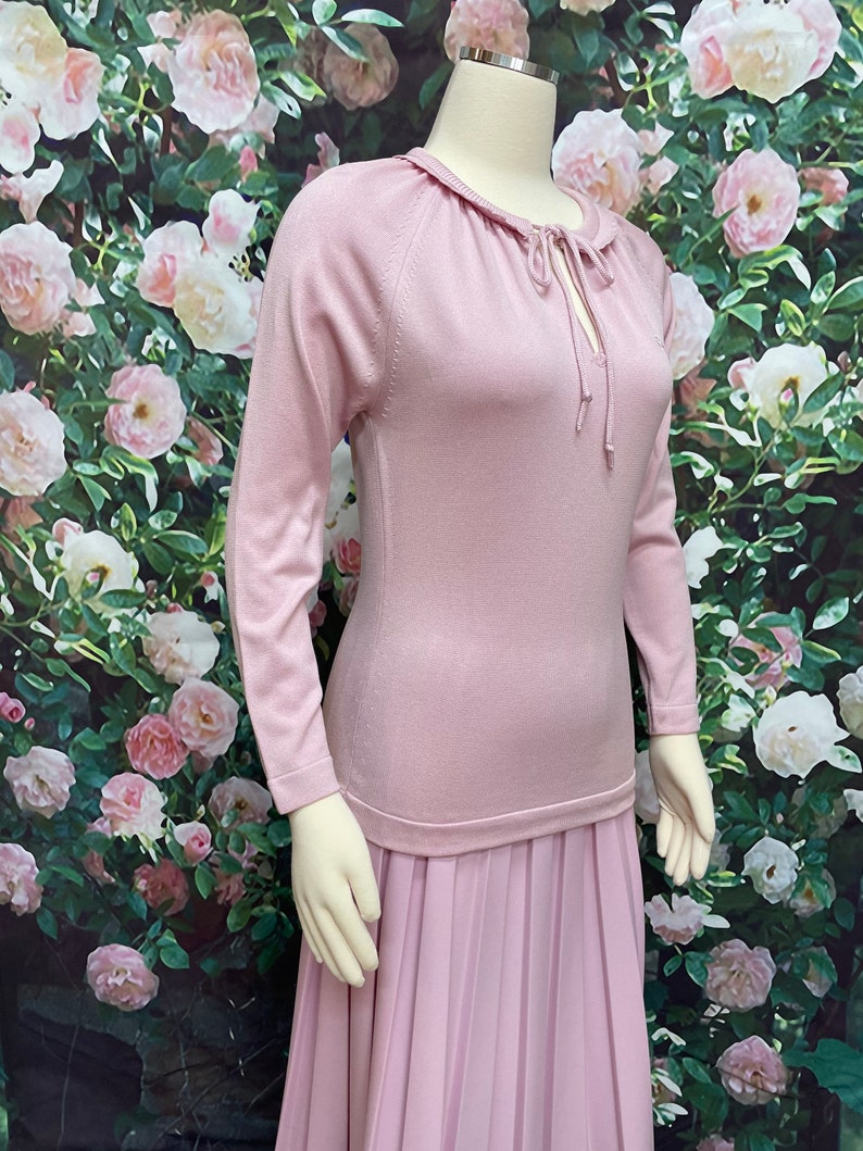 May include: A long, pink dress with a pleated skirt and a fitted bodice. The bodice has a round neckline with a tie detail.