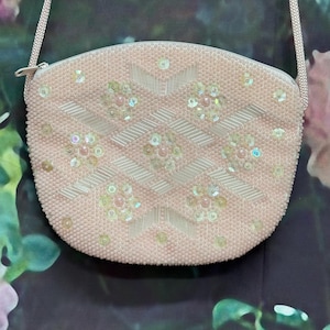 May include: A pale pink beaded handbag featuring a geometric pattern of clear iridescent sequins and elongated glass beads. The purse has a rounded shape, a top zipper closure, and a matching thin shoulder strap made of woven fabric.
