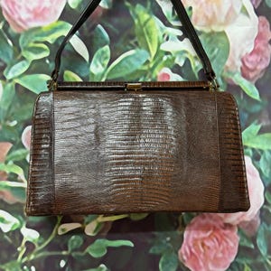 May include: A vintage brown handbag with a textured, reptile-like pattern. The rectangular bag features a clasp closure and a single handle. The background is a floral pattern with pink and green hues.