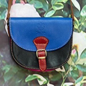 Peut inclure: Un sac à bandoulière de style selle avec un rabat en cuir bleu, un corps en cuir noir et une sangle en cuir rouge avec une boucle en métal doré. Le rabat bleu est gaufré d'un emblème fleur de lys et du texte Made in Italy.