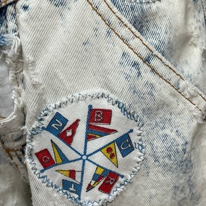 Puede incluir: Una chaqueta vaquera blanca con un parche azul y rojo con un dise&ntilde;o de bandera n&aacute;utica. El parche est&aacute; cosido a la chaqueta con una puntada en zigzag.
