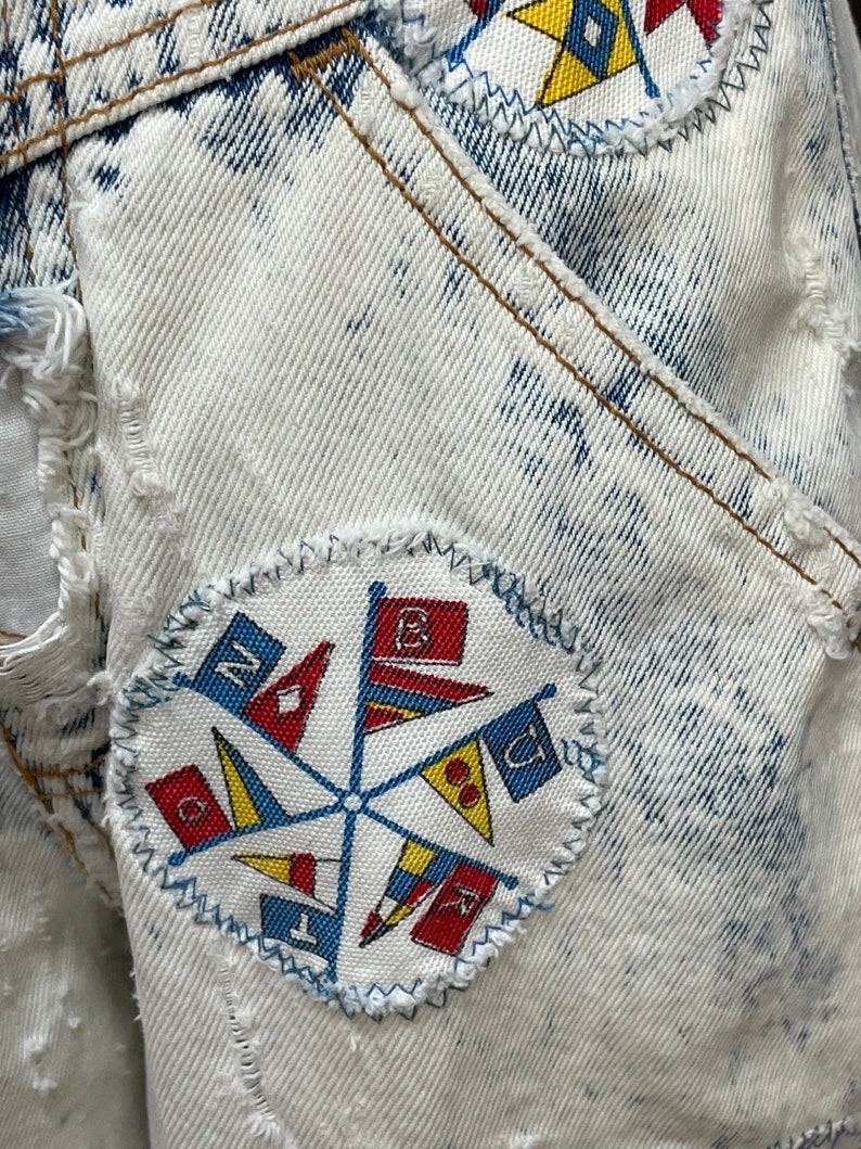 Puede incluir: Una chaqueta vaquera blanca con un parche azul y rojo con un dise&ntilde;o de bandera n&aacute;utica. El parche est&aacute; cosido a la chaqueta con una puntada en zigzag.