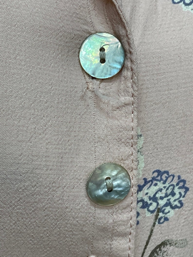 May include: Two pearly white buttons with a light brown thread running through the center holes. The buttons are sewn onto a light pink fabric with a floral pattern.