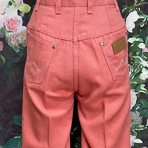 May include: A pair of wide-leg, light pink denim pants with a white stitching detail and a brown leather patch with the word "Wrangler" on the back right pocket.
