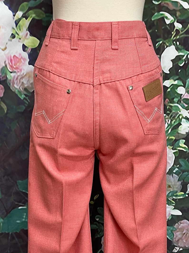 May include: A pair of wide-leg, light pink denim pants with a white stitching detail and a brown leather patch with the word "Wrangler" on the back right pocket.