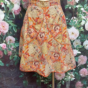 May include: A vintage, knee-length skirt with a patchwork design in shades of orange, yellow, and red. The skirt features a geometric pattern with floral accents and a scalloped orange trim along the hemline. The skirt is displayed on a mannequin.