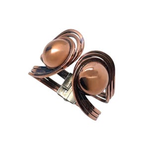 May include: A copper-toned cuff bracelet with a unique design. The bracelet features two large, spherical accents and a series of curved, layered metal bands. A silver-colored clasp secures the bracelet. The overall aesthetic is modern and artistic.