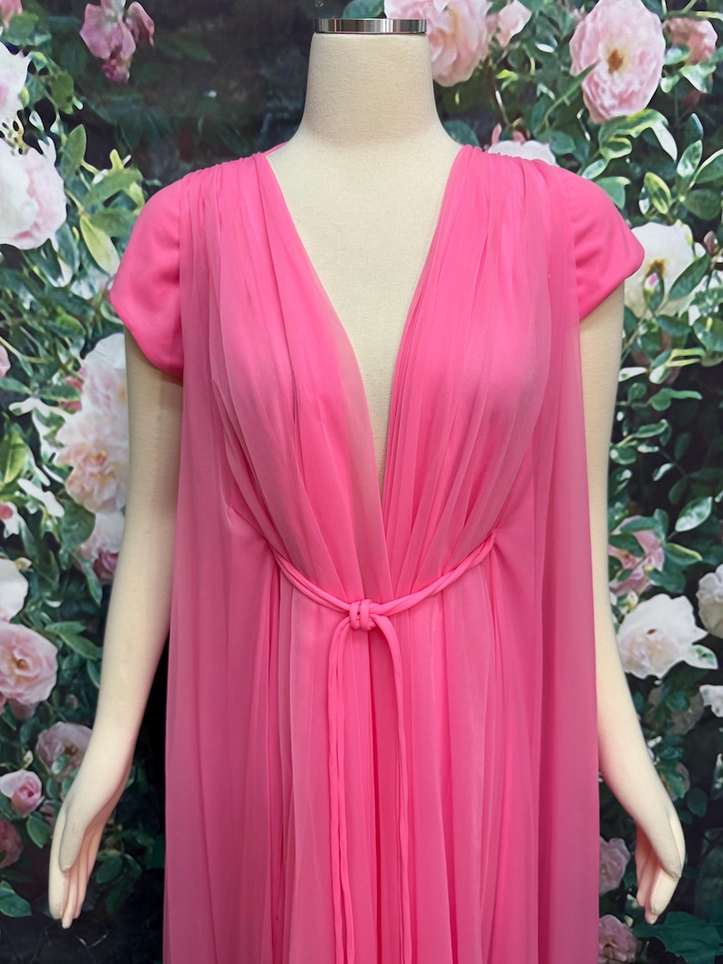 May include: A bright pink floor-length gown featuring a deep V-neckline, short sleeves, and a sheer, layered chiffon overlay. The garment includes a matching fabric tie at the waist, creating a gathered effect across the front bodice.