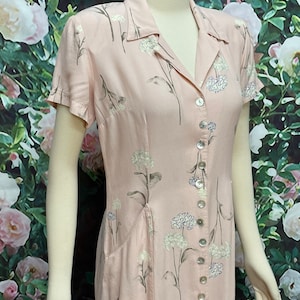 May include: A pink floral button-down dress with short sleeves and a single pocket on the left side. The dress has a white floral pattern and a button closure down the front.