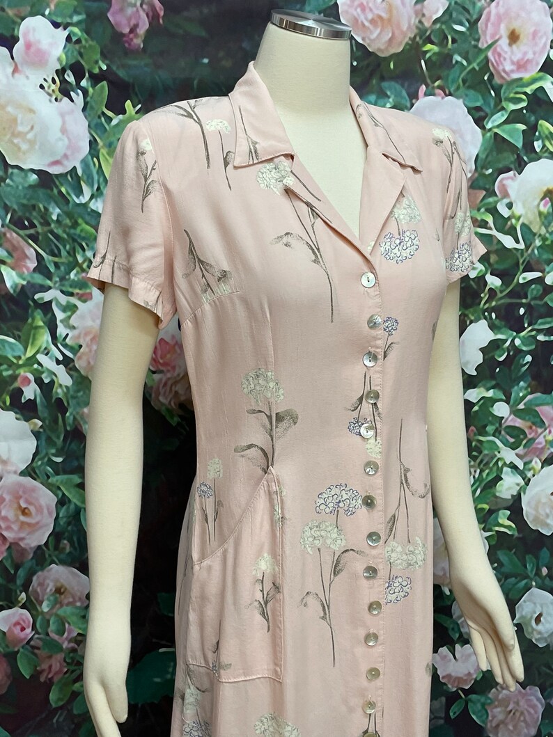 May include: A pink floral button-down dress with short sleeves and a single pocket on the left side. The dress has a white floral pattern and a button closure down the front.