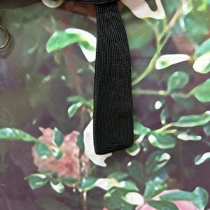 May include: A black fabric strap with a woven texture. The strap is against a background of a floral print with green leaves and pink flowers.