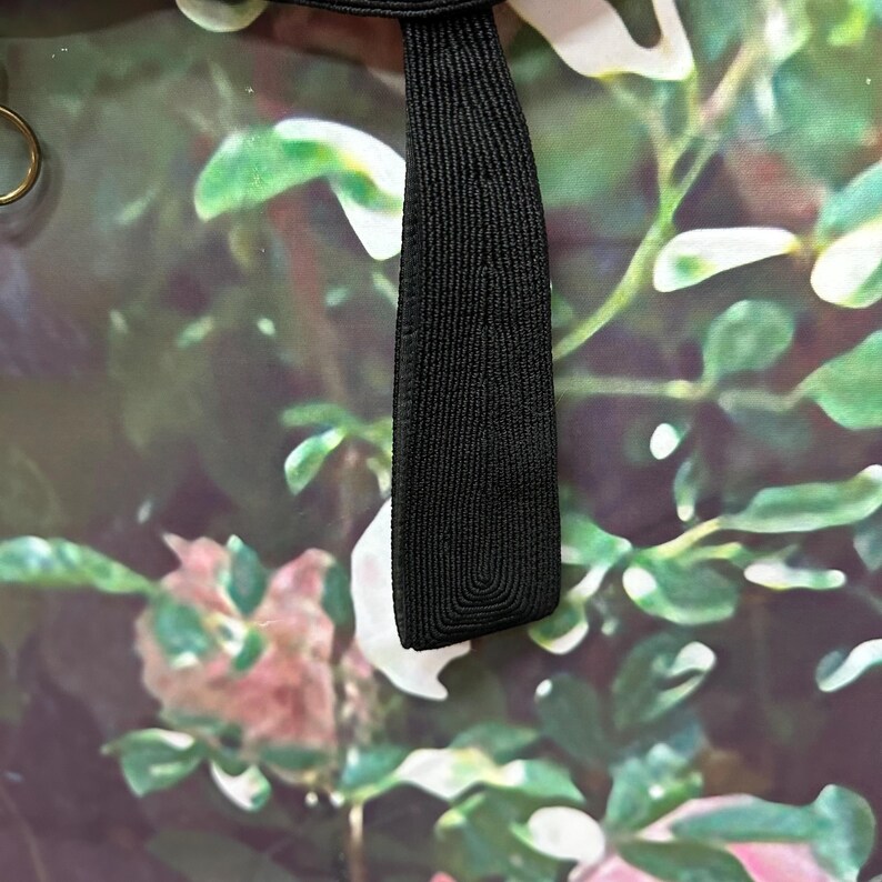 May include: A black fabric strap with a woven texture. The strap is against a background of a floral print with green leaves and pink flowers.