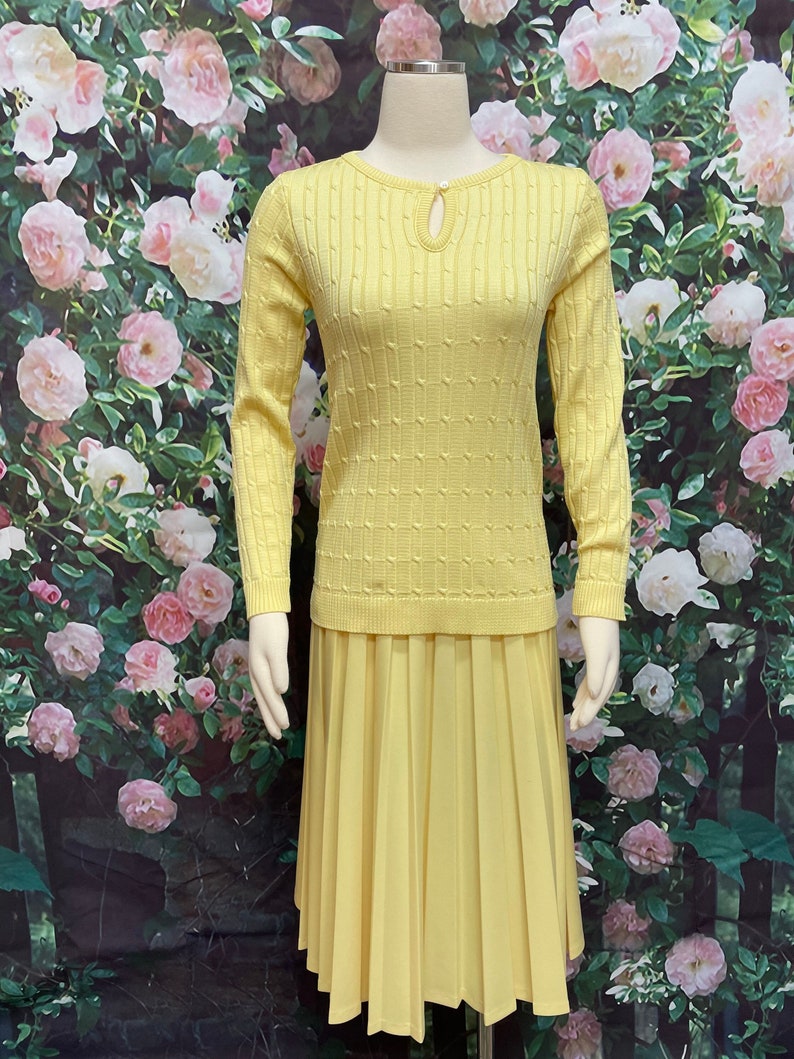 May include: A yellow knitted sweater with a keyhole neckline and a matching pleated skirt. The sweater has a textured pattern and the skirt is knee-length.