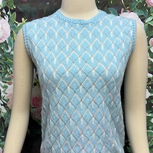 May include: A sleeveless knit top in light blue featuring a repeating diamond pattern with white accents. The garment has a round neckline and a textured border along the armholes and hem, showcasing a delicate, openwork stitch design.