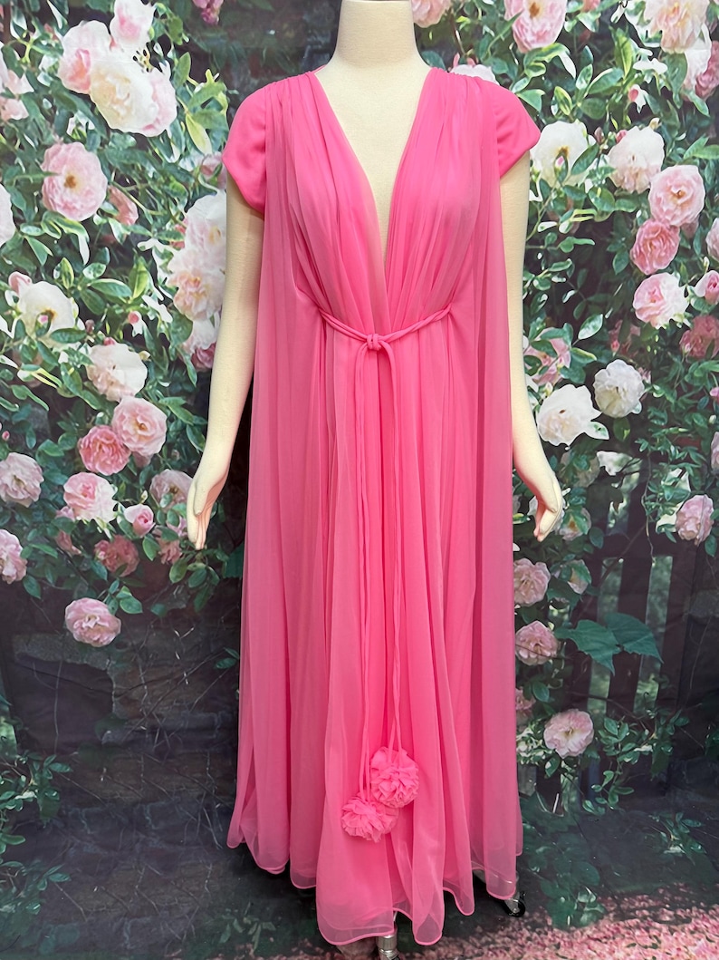 May include: A floor-length, bright pink gown featuring a deep V-neckline, short sleeves, and a sheer, flowing overlay. The garment includes a thin, matching pink tie at the waist that secures with two decorative fabric flower accents at the ends.