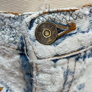 Puede incluir: Primer plano de un jean de mezclilla desgastado con un bot&oacute;n de metal que dice "ZENA" en may&uacute;sculas.
