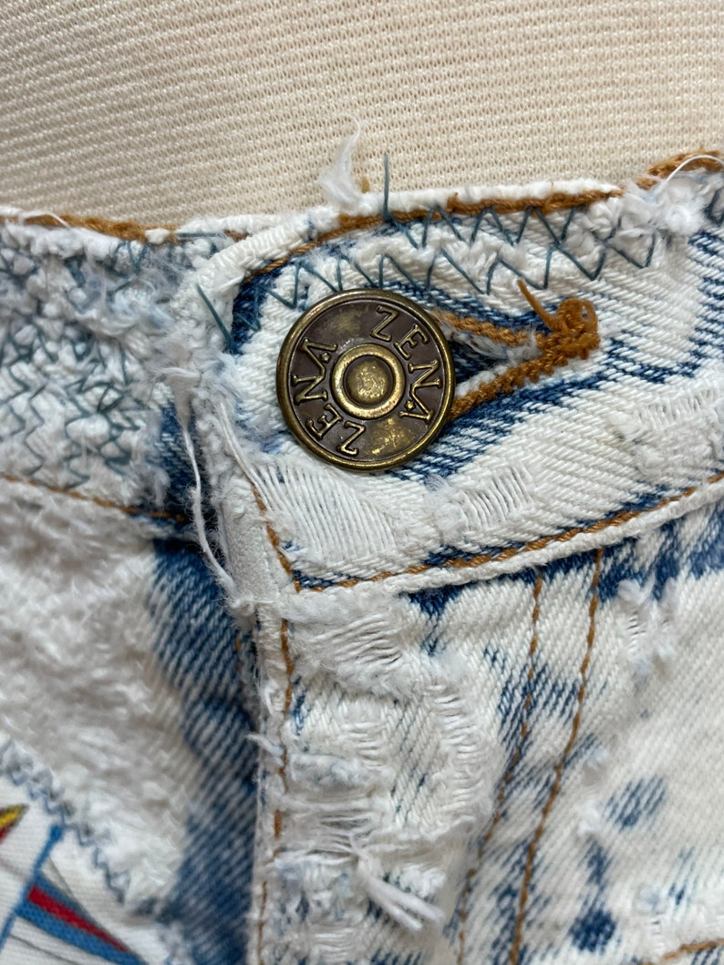 Puede incluir: Primer plano de un jean de mezclilla desgastado con un bot&oacute;n de metal que dice "ZENA" en may&uacute;sculas.