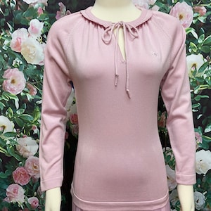 May include: A pale pink long-sleeved sweater with a ruffled neckline and a tie at the neck. The sweater has a small embroidered logo on the right side of the chest.