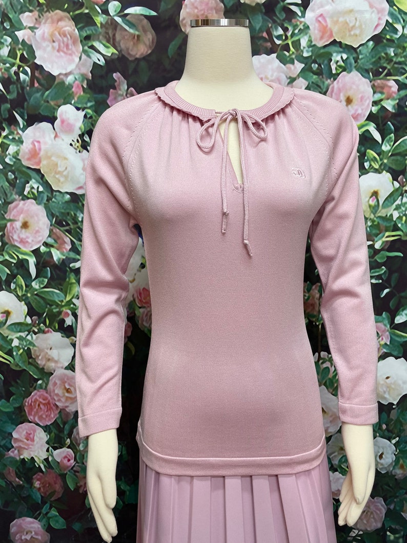 May include: A pale pink long-sleeved sweater with a ruffled neckline and a tie at the neck. The sweater has a small embroidered logo on the right side of the chest.