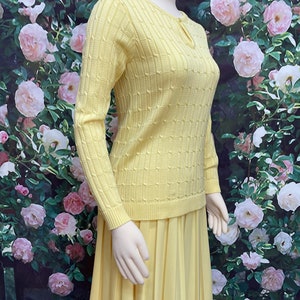 May include: A yellow knitted sweater with a keyhole neckline and a matching pleated skirt. The sweater has a cable knit pattern and the skirt is made of a smooth fabric.