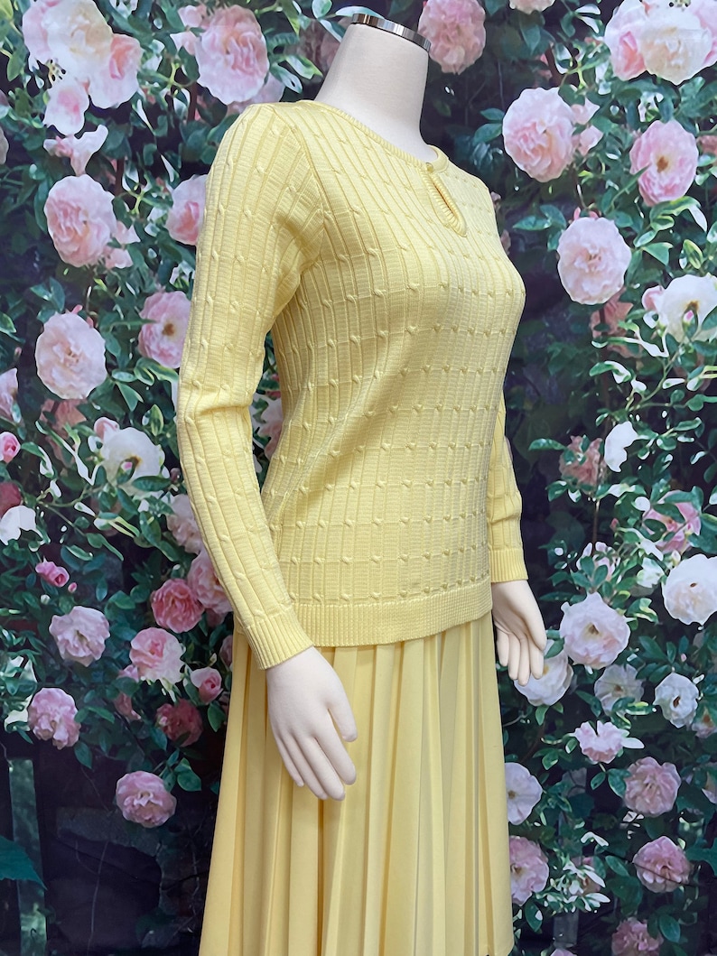 May include: A yellow knitted sweater with a keyhole neckline and a matching pleated skirt. The sweater has a cable knit pattern and the skirt is made of a smooth fabric.