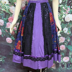 May include: A purple skirt with a floral patterned fabric, black lace, and a purple hem. The skirt has a wide waistband and is displayed on a mannequin.