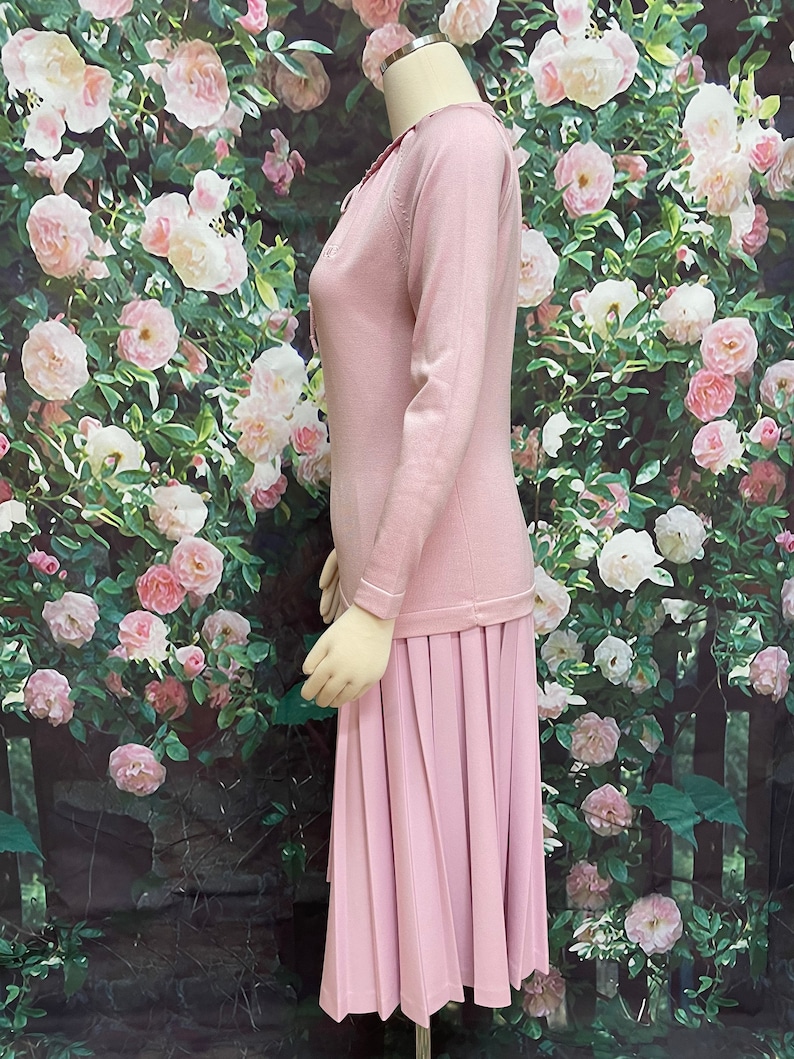 May include: A pink knitted dress with a pleated skirt. The dress has a round neckline and long sleeves.
