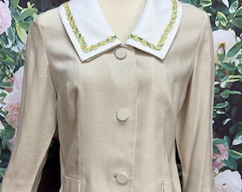 May include: A beige blazer with a white collar and three buttons. The collar and sleeve cuffs are trimmed with a yellow and green floral design. The blazer has three-quarter length sleeves and two front pockets.