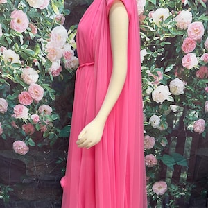 May include: A floor-length, bright pink sleeveless dress featuring a sheer, flowing overlay that drapes from the shoulders. The garment includes a thin, matching pink tie belt at the waist, creating a gathered effect along the bodice and skirt.