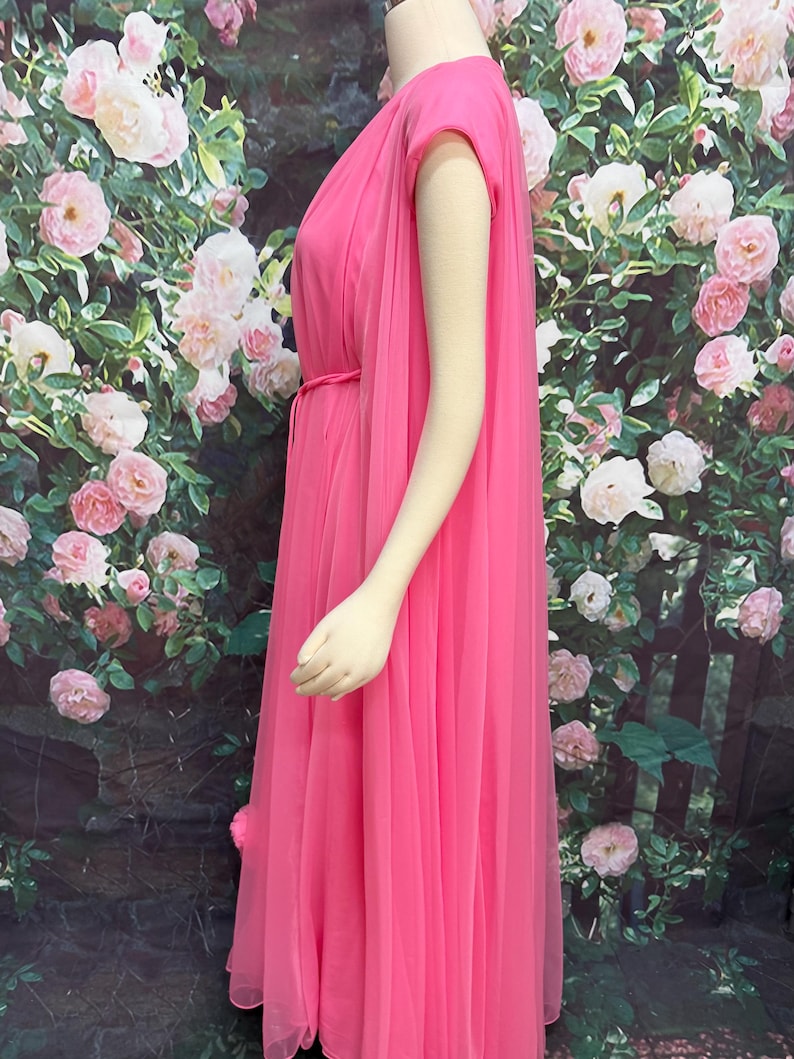 May include: A floor-length, bright pink sleeveless dress featuring a sheer, flowing overlay that drapes from the shoulders. The garment includes a thin, matching pink tie belt at the waist, creating a gathered effect along the bodice and skirt.