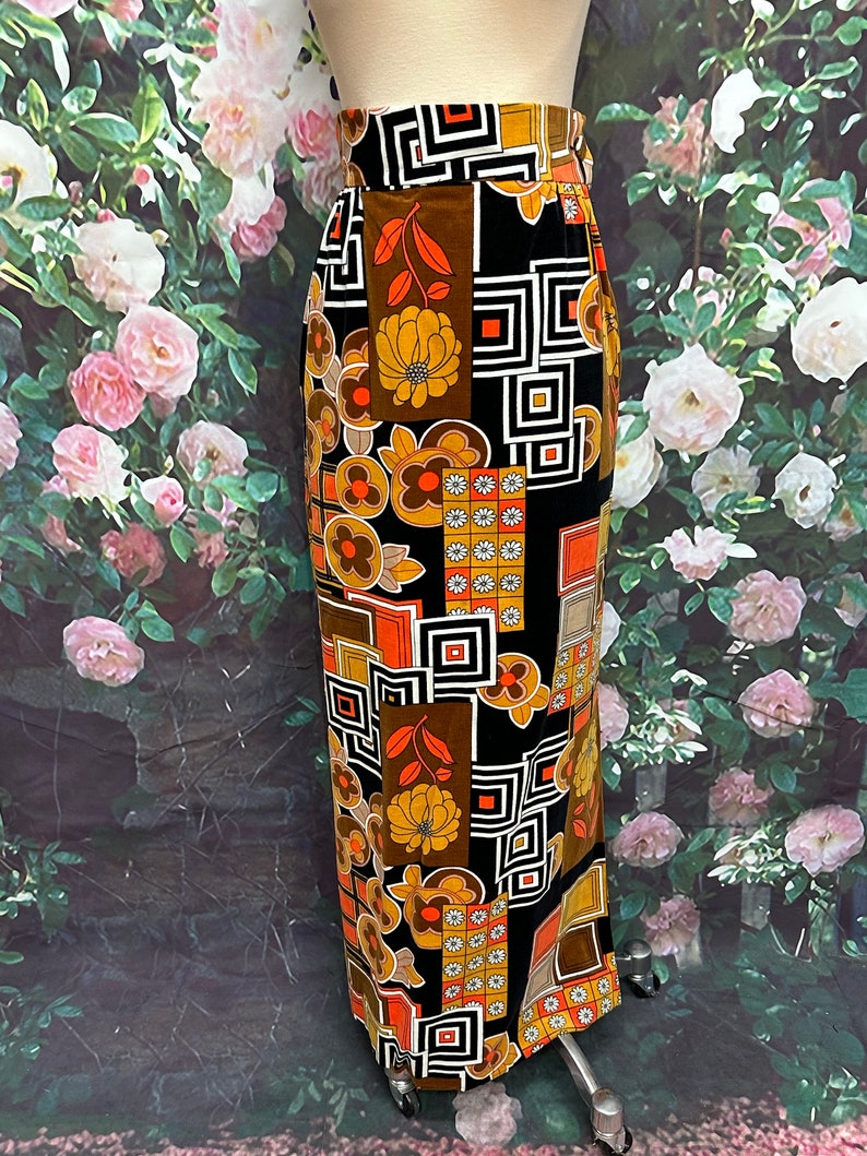 May include: A long black skirt with a geometric and floral print in shades of orange, brown, and white. The skirt has a high waist and a straight silhouette.