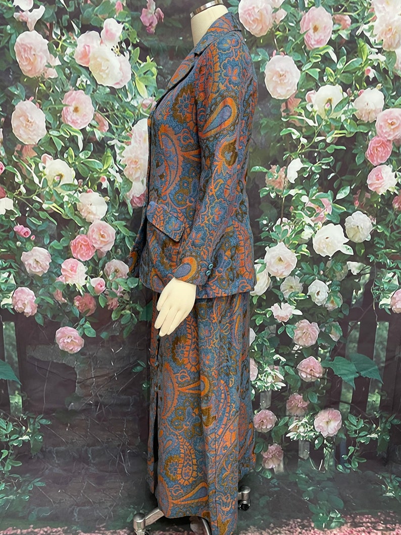 May include: A blue and orange paisley patterned skirt suit. The skirt is long and has a slit up the front. The jacket has a collar and long sleeves.
