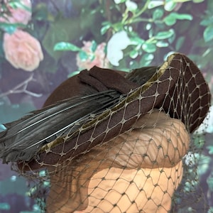 May include: A dark brown felt hat featuring a structured brim with a thin olive green velvet trim. The hat is adorned with a cluster of long, dark feathers and a delicate, fine-mesh netting that drapes over the front of the headpiece.