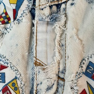 Puede incluir: Primer plano de un par de pantalones vaqueros de mezclilla azul claro con un aspecto desgastado. Los vaqueros tienen parches de tela cosidos, con un tema n&aacute;utico con banderas rojas, amarillas y azules. Los vaqueros tienen un borde deshilachado y una costura rasgada.