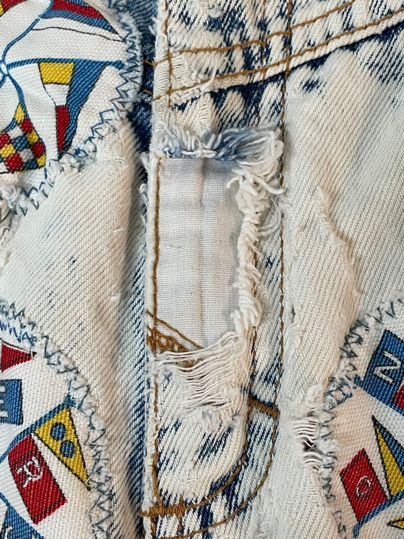 Puede incluir: Primer plano de un par de pantalones vaqueros de mezclilla azul claro con un aspecto desgastado. Los vaqueros tienen parches de tela cosidos, con un tema n&aacute;utico con banderas rojas, amarillas y azules. Los vaqueros tienen un borde deshilachado y una costura rasgada.