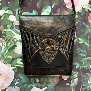 May include: Black leather shoulder bag with gold and snakeskin accents. The bag features a decorative flap with a geometric design and a gold-colored embellishment. The bag has a long strap for over-the-shoulder wear.
