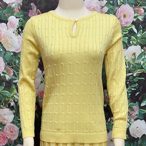 May include: A yellow knitted sweater with a keyhole neckline and a textured pattern. The sweater has long sleeves and a relaxed fit.