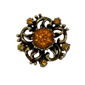 Pu&ograve; includere: Un fermaglio dorato con un grande strass color ambra al centro. Il fermaglio &egrave; decorato con strass pi&ugrave; piccoli e un design a spirale ornato.