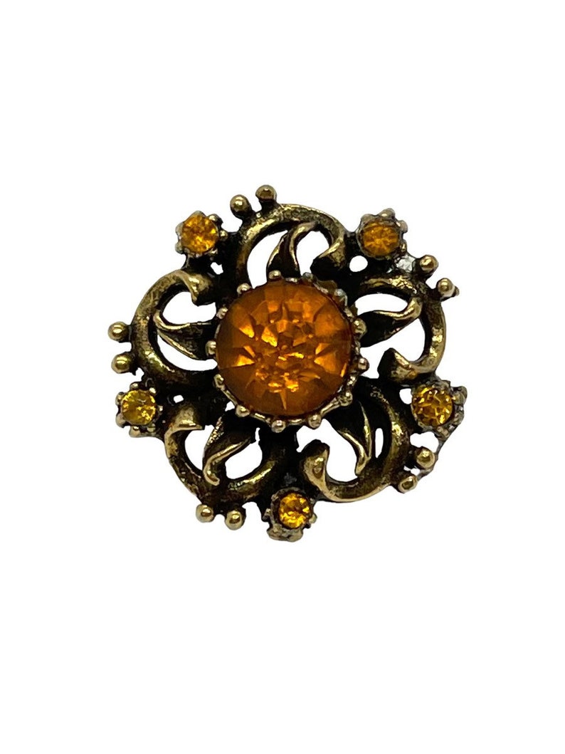 Pu&ograve; includere: Un fermaglio dorato con un grande strass color ambra al centro. Il fermaglio &egrave; decorato con strass pi&ugrave; piccoli e un design a spirale ornato.