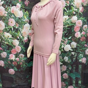 May include: A pink sweater dress with a pleated skirt and a small embroidered logo on the chest. The dress has a drawstring neckline and long sleeves.