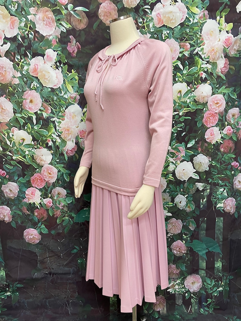 May include: A pink sweater dress with a pleated skirt and a small embroidered logo on the chest. The dress has a drawstring neckline and long sleeves.