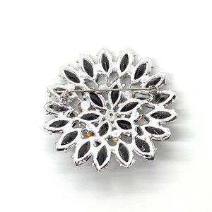 May include: A silver brooch with a circular design featuring black and clear rhinestones. The brooch has a pin on the back for attaching to clothing.