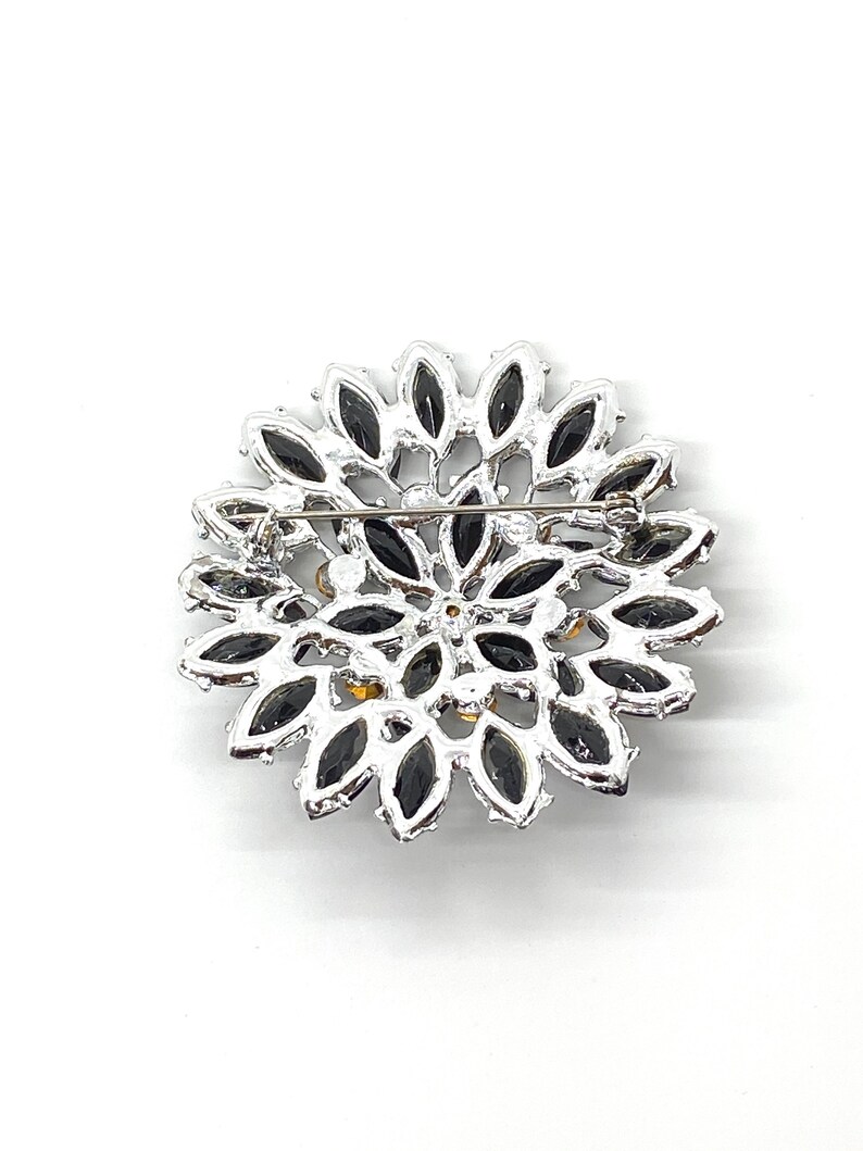 May include: A silver brooch with a circular design featuring black and clear rhinestones. The brooch has a pin on the back for attaching to clothing.