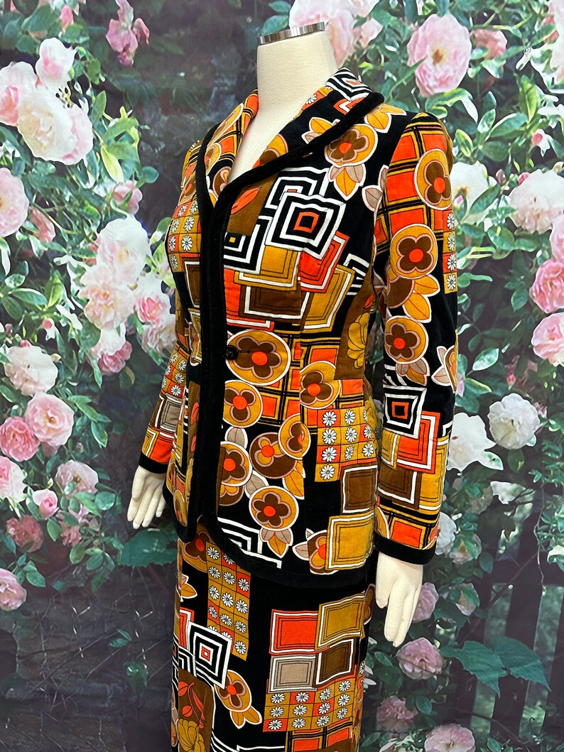 May include: A black and brown vintage skirt suit with a geometric and floral pattern. The suit features a fitted jacket with a collar and a skirt with a straight silhouette.