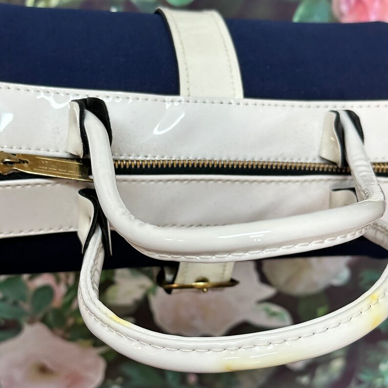 May include: A white and navy blue handbag with a white patent leather finish. The bag has two top handles and a zipper closure.